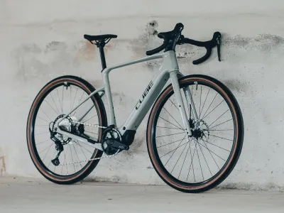 Cube Nuroad Hybrid C:62 Race 400X in reedbeige´n´black (Beige/Schwarz) – Schrägansicht von vorne rechts, an Betonwand gelehnt